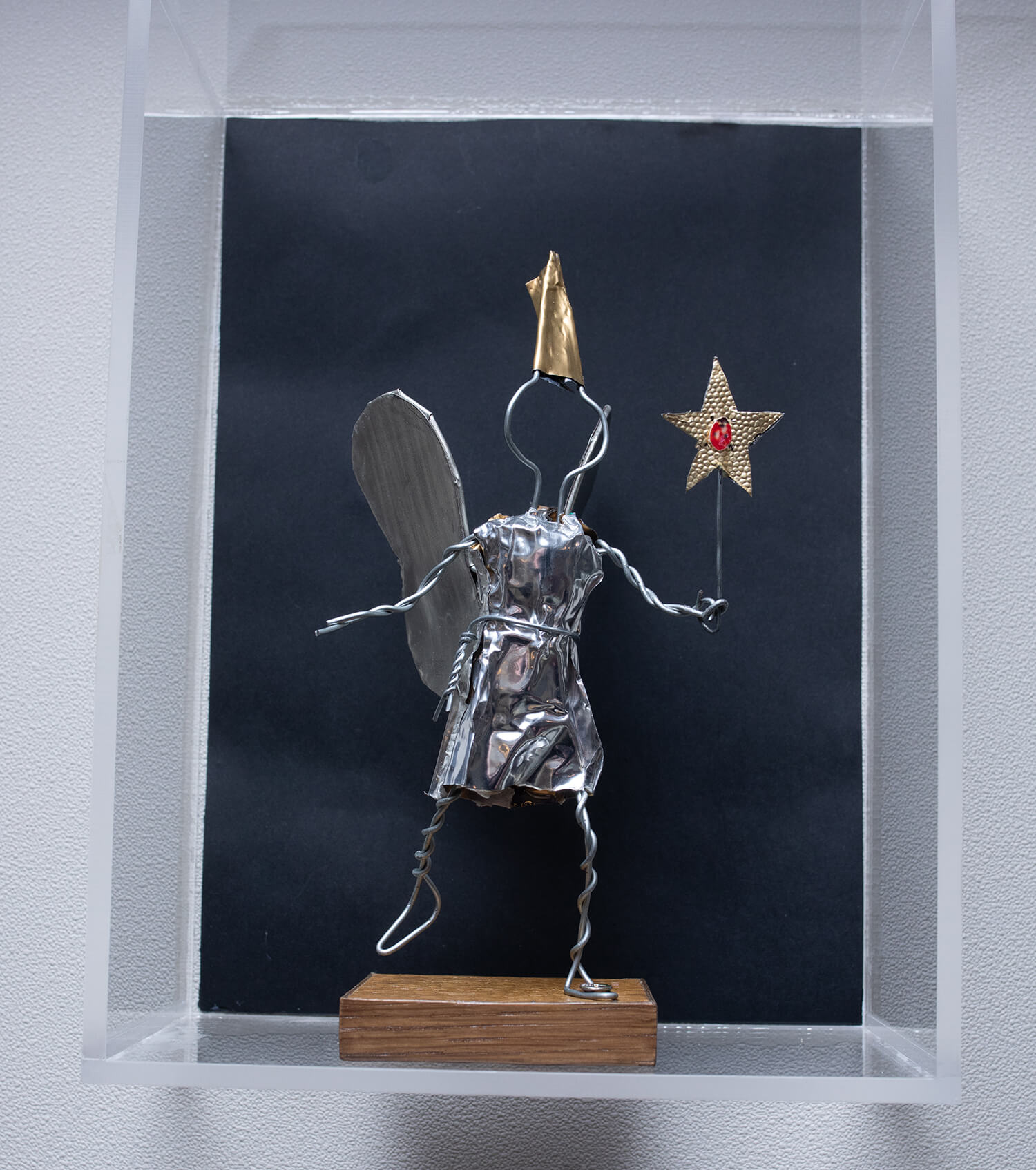 On voit la petite figurine de la Fée Bulléo sur un socle rectangulaire en bois, faite à partir du fil de fer torsadé du muselet et de la collerette argentée de bouchon de champagne. Elle tient une baguette magique en forme d’étoile dorée avec une coccinelle au centre. Elle a des ailes argentées et un chapeau pointu doré.