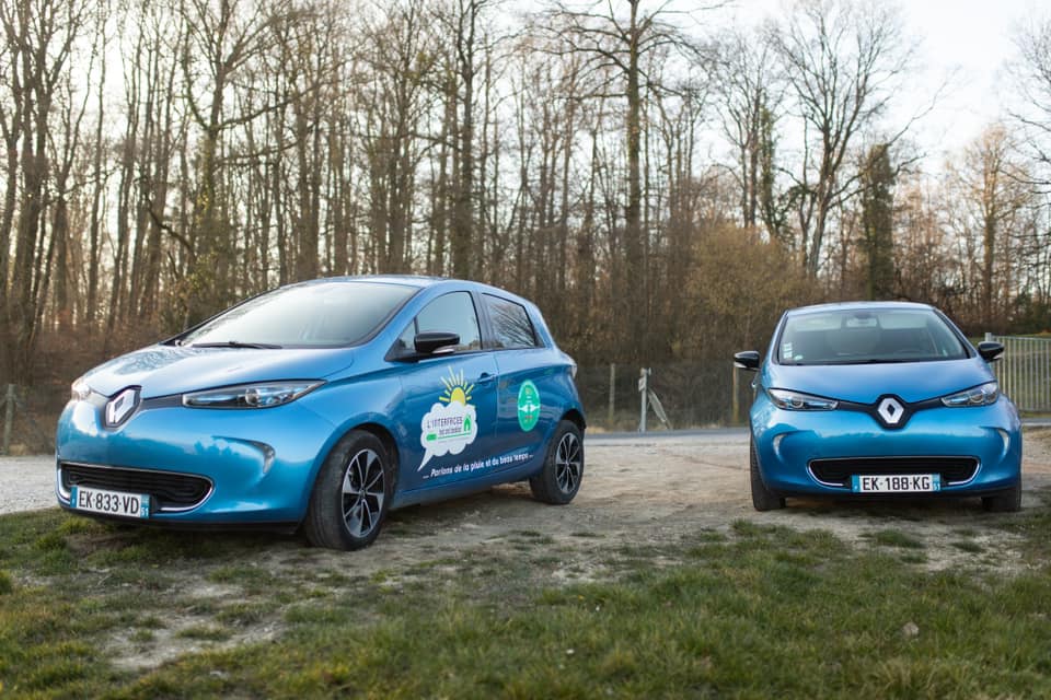 On voit les 2 Renault Zoé électriques garées dans un chemin de terre devant des arbres sur le coté d’une route