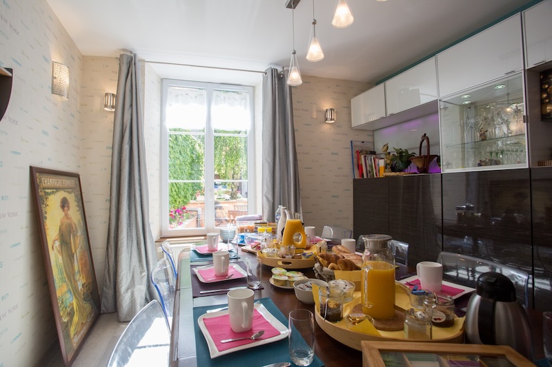 On voit la table du petit-déjeuner dressée dans la salle à manger de Linterfaces, garnie de viennoiseries et baguettes du boulanger, produits faits maison, fruits et jus de fruits et bien d’autres choses sucrées et salées.